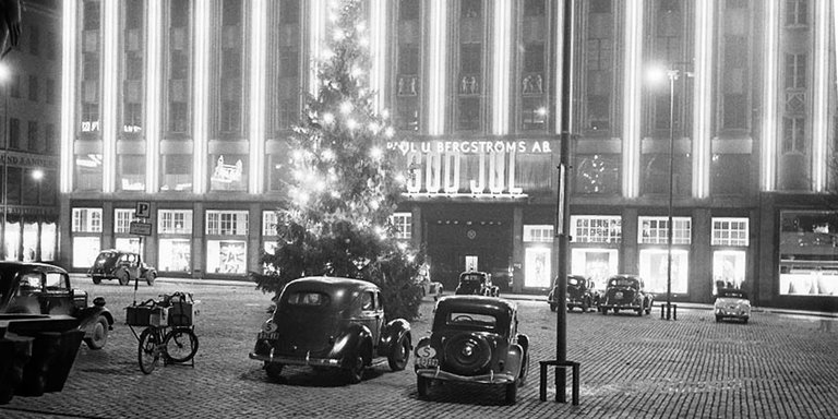 Old black and white photo of the PUB-department store, taken at Hötorget in Stockholm. Nightime. Old cars are parked at the square in front of the department store, the exterior is lit up by bright lights. Greta Garbo was discovered at the up-scale department store before moving to Hollywood.