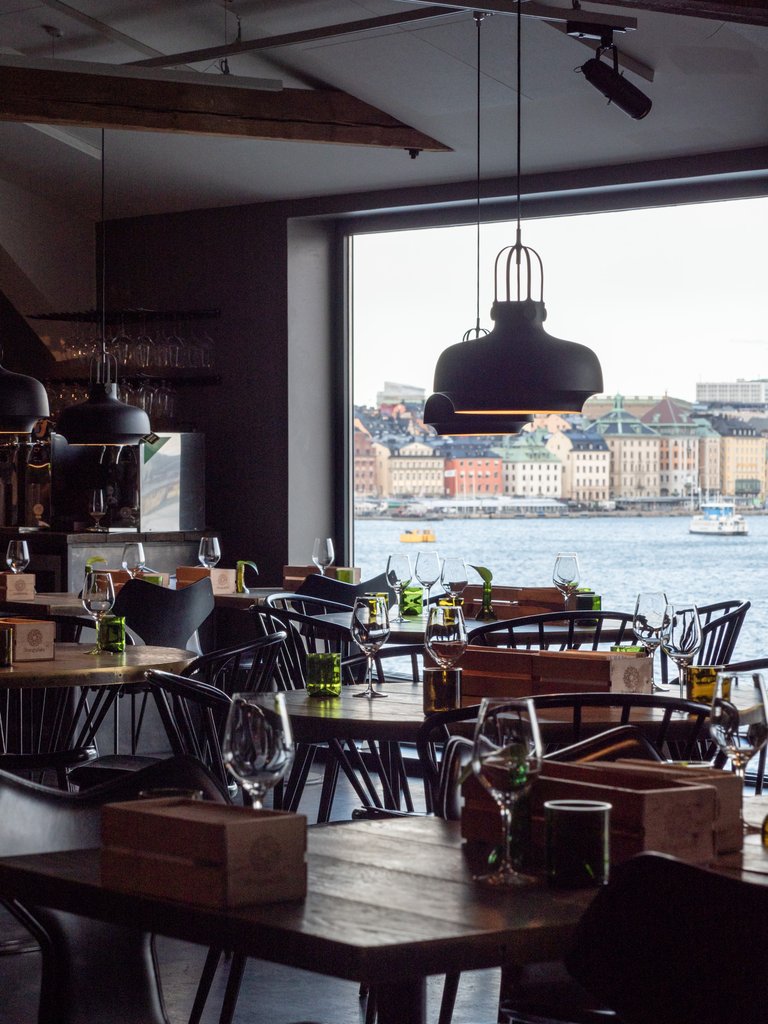 Restauranginteriör med dukade träbord och pendellampor, med utsikt över vattnet och Stockholm genom ett stort fönster.