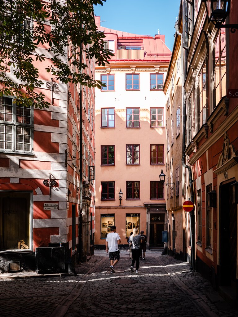 Smal kullerstensgränd i Gamla stan i Stockholm, med färgglada fasader i rött och rosa och människor som promenerar i solen.