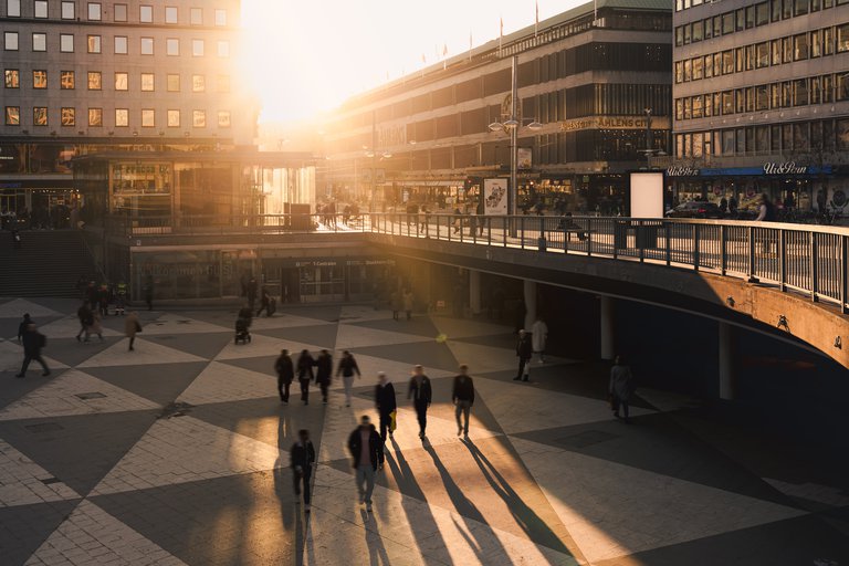 Vår i Stockholm. Solen går ner över Sergels Torg. Människor går över det centralt belägna torget medans solens strålar syns mellan byggnaderna.