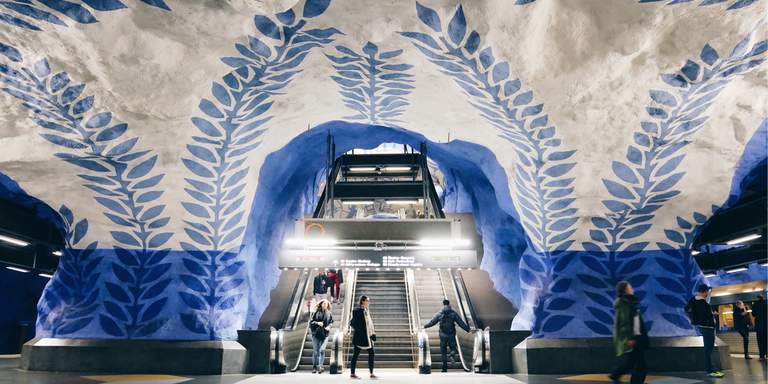 T-Centralen subway station in Stockholm. The blue line platform. A cave painted in blue and white. People ride down an escalator to the platform.