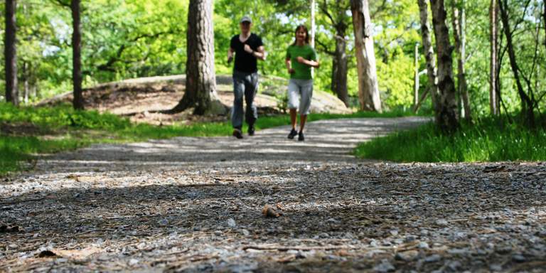 Sommar i Stockholm. En man och en kvinna joggar mot kameran längs med en skogsstig i Nacka naturreservat.