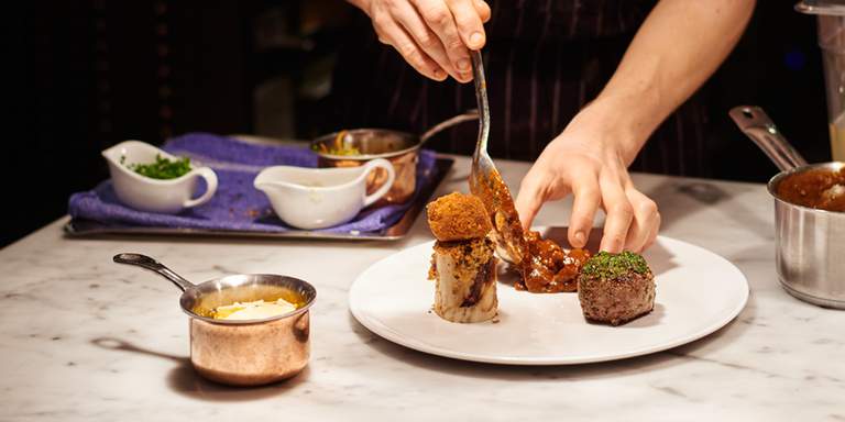 Ett par händer förbereder mat till en middagsservering på restaurang Luzette. Ljuset är dunkelt och personen på bilden gör de sista avslutade detaljarbetet med maträtten innan den skickas ut. 