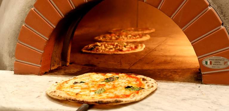 En traditionell italiensk pizza med tunna kanter är på väg in i en öppen vedugn. Två till pizzor är redan inne i ugnen och ser supergoda ut.