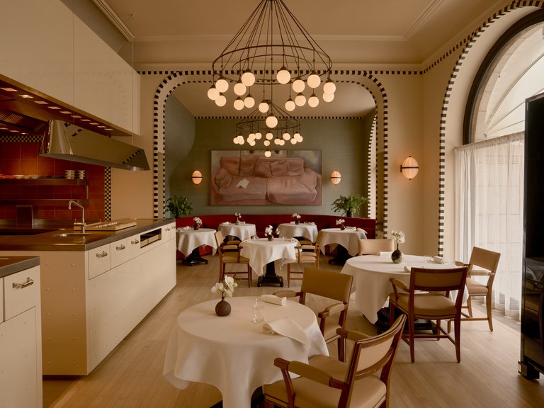 Elegant restauranginteriör med vitdukade bord, öppet kök, höga välvda fönster och dekorativa ljuskronor.