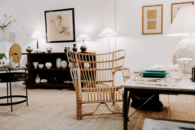 Inredd monter med antika möbler, rottingfåtölj, bordslampor och konstverk i en mässhall.