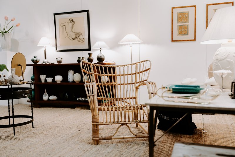 Inredd monter med antika möbler, rottingfåtölj, bordslampor och konstverk i en mässhall.