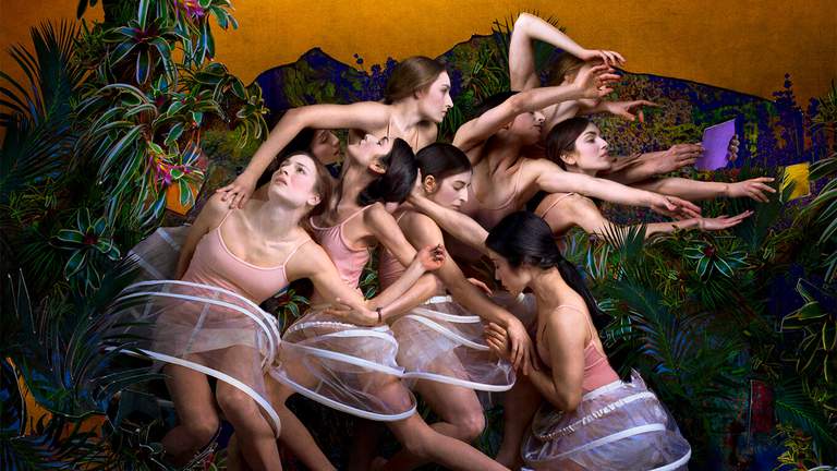 En grupp unga kvinnor i underkläder står sammanpackade i en djungelmiljö. De tittar åt olika håll och sträcker sig efter olika saker likt en renässansmålning.