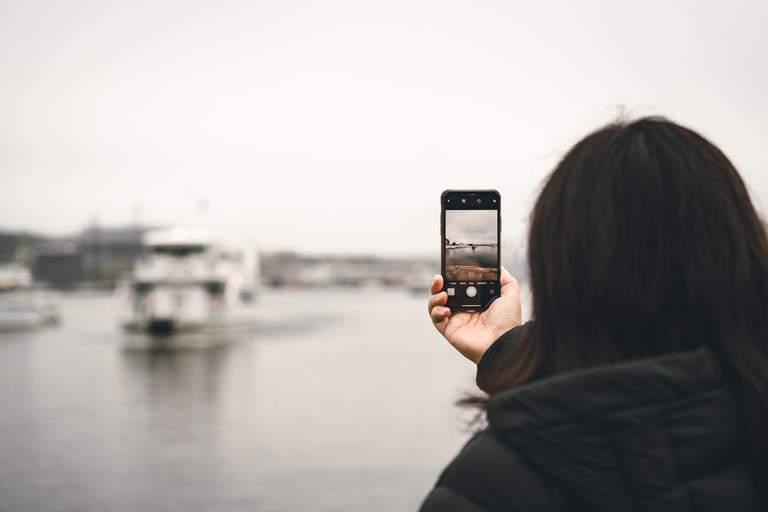 En person håller upp en mobiltelefon för att ta en bild på en vit skärgårdsfärja som kör i vattnet i centrala Stockholm