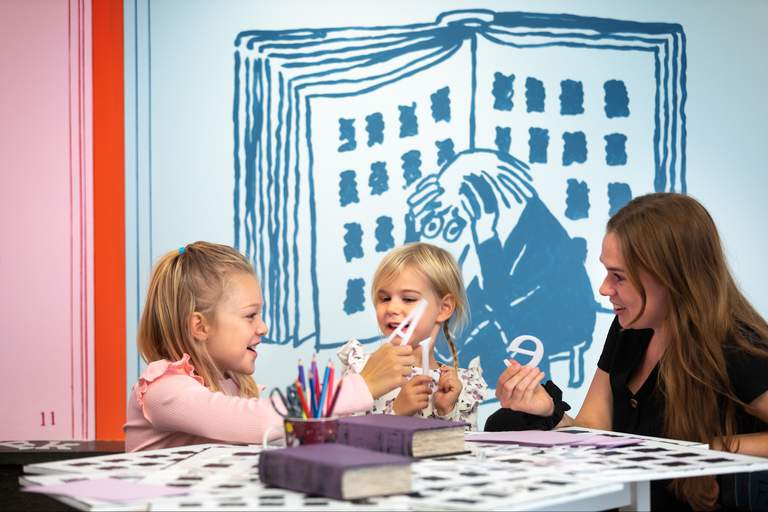 En kvinna och två barn sitter vid bordet med böcker, pennor, papper och andra föremål på det.