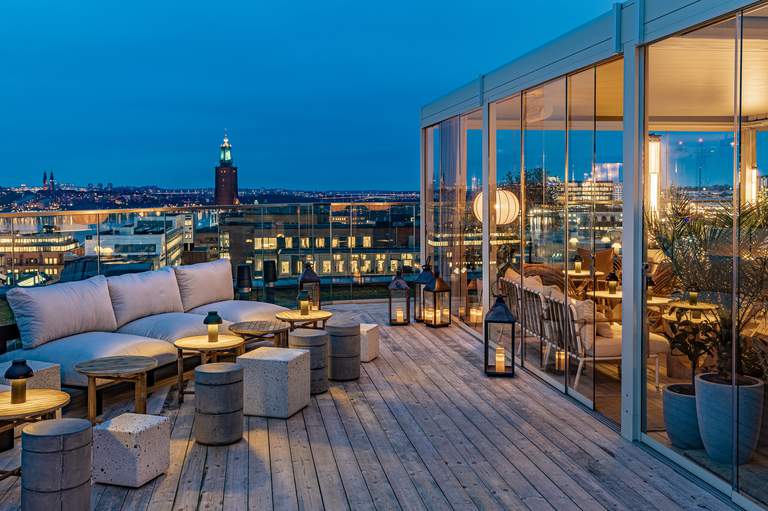 En takterrass i skymningen. Ljusa soffor och låga soffbord med ljus som skiner i mörkret. Stockholms Stadshus syns i bakgrunden.