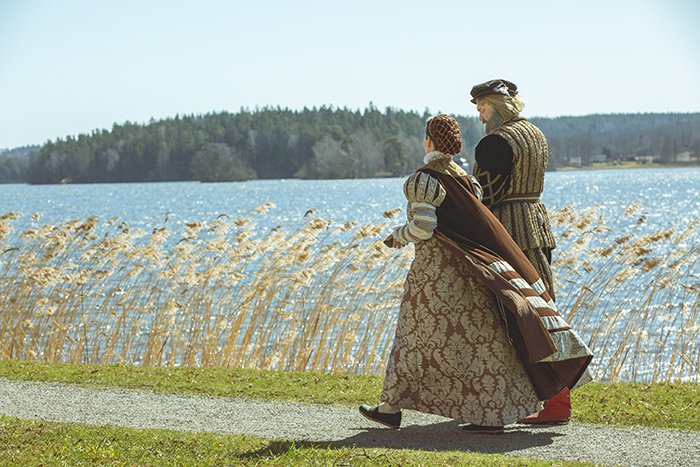 Två personer i historiska dräkter som promenerar på en sjöstrand.