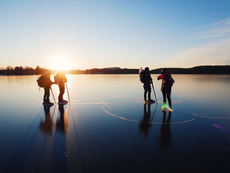 Aktiviteter i Stockholm. Vinter. Fyra personer åker långfärdsskridskor. På bilden har de stannat mitt på en sjö, med solnedgång vid horisonten.