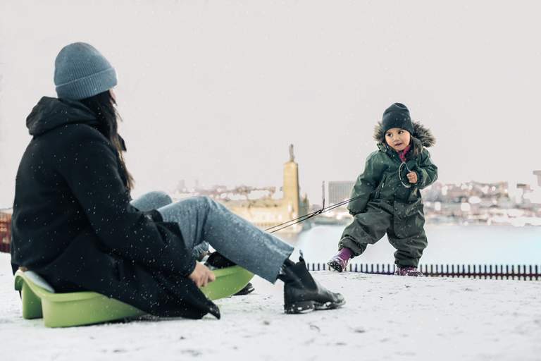 Ett barn i overall och mössa drar sin förälder i en pulka i snön. Det är snöflingor i luften och i bakgrunden syns Stockholms Stadshus.