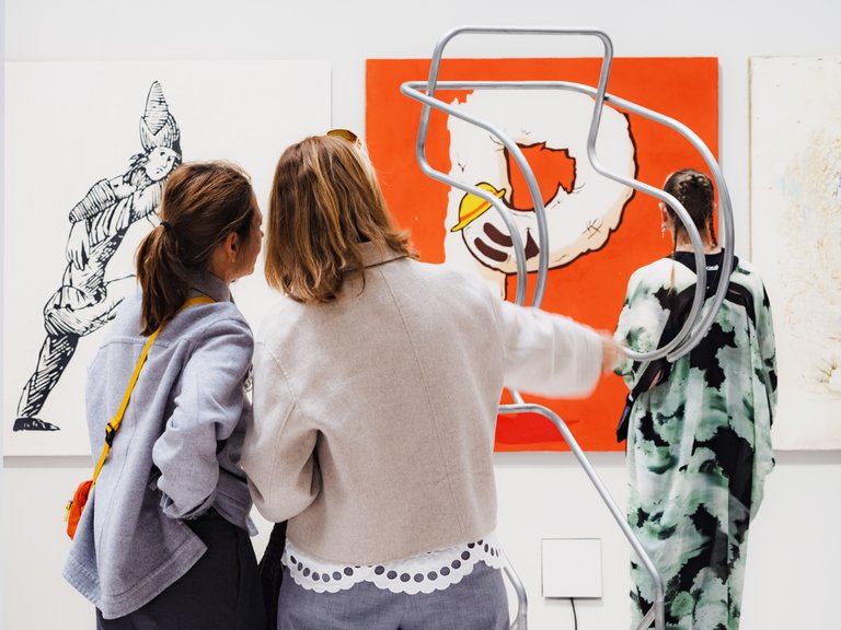 Besökare på en konstmässa tittar på målningar och en skulptural metallform i ett galleri.