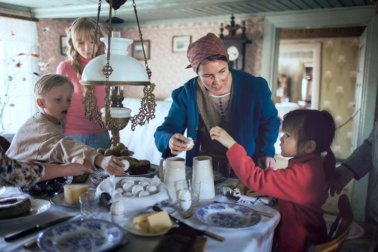 en kvinna och tre barn står vid ett bord.  på bordet finns tallrikar, ägg, smör och andra föremål