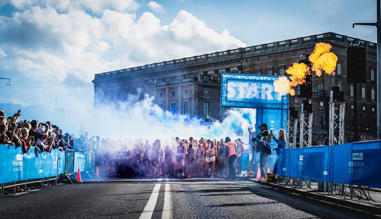 En klunga med löpare springer mot kameran då starten just gått i Stockholms Halvmarathon, precis framför kungliga slottet, en solig dag i september.