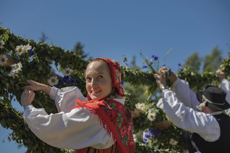 Kvinna i traditionell folkdräkt dekorerar en midsommarstång med gröna kvistar och blommor, tillsammans med andra personer i bakgrunden. Hon ler mot kameran under en klarblå himmel.