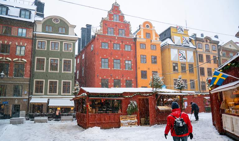 Röda marknadsbås dekorerade med lampor och granris står på ett snöigt torg kantat av färgglada, historiska byggnader