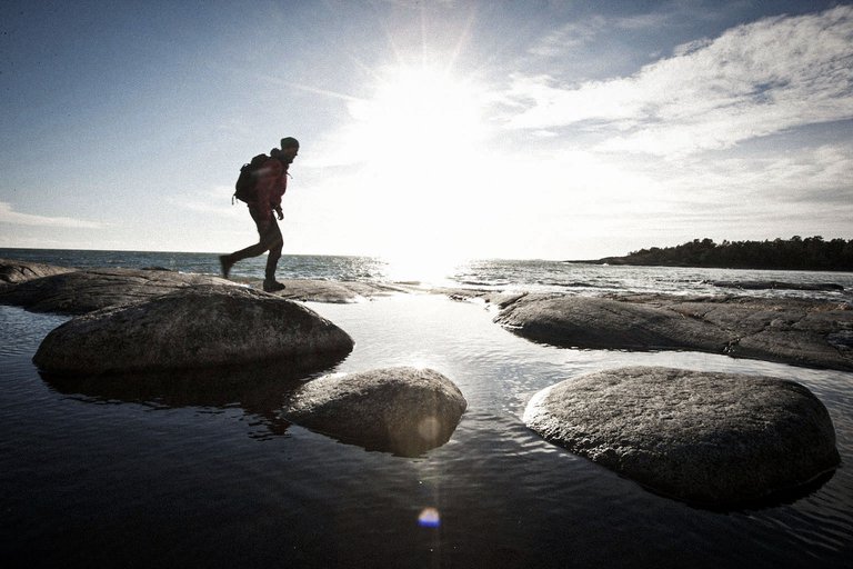 En vandrare tar steg över släta havsnära klippor, motljus av den starka solen nära horisonten.