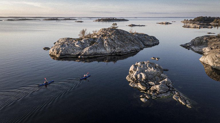 Två kajakpaddlare glider fram mellan karga, trädtoppade öar i lugnt vatten under en mjuk kvällshimmel.