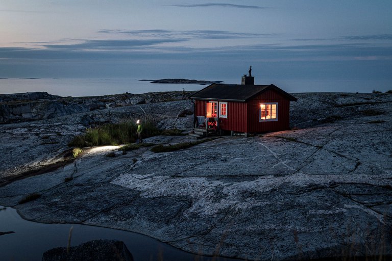 En mysig röd stuga med tända fönster ligger på en klippig strandlinje i skymningen, ett stilla hav i bakgrunden.
