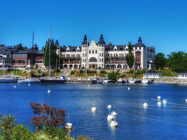 Byggnaden av Grand Hotel i Saltsjöbaden nära vattnet med flera segelbåtar vid kajen.