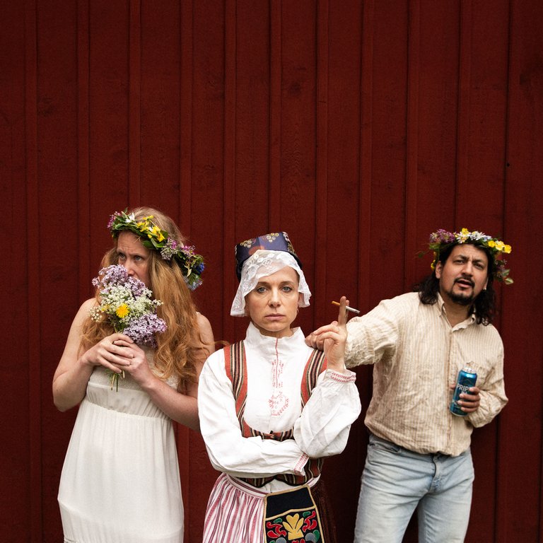 Tre personer i midsommar-utstyrsel står mot en röd vägg.