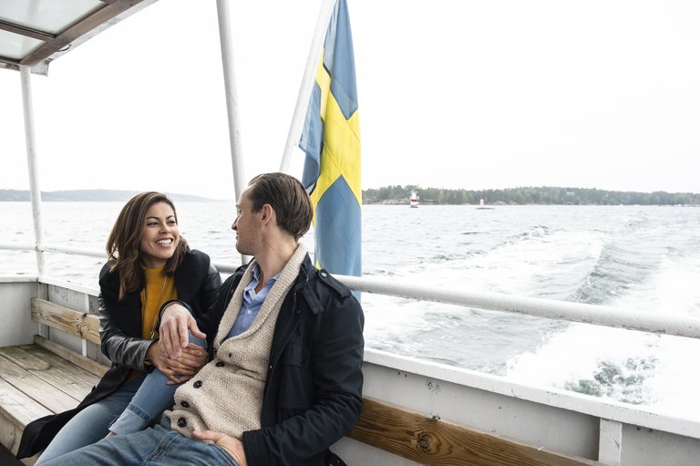Ett par sitter på däck på en skärgårdsbåt och ler mot varandra, med svensk flagga och öppet vatten i bakgrunden.