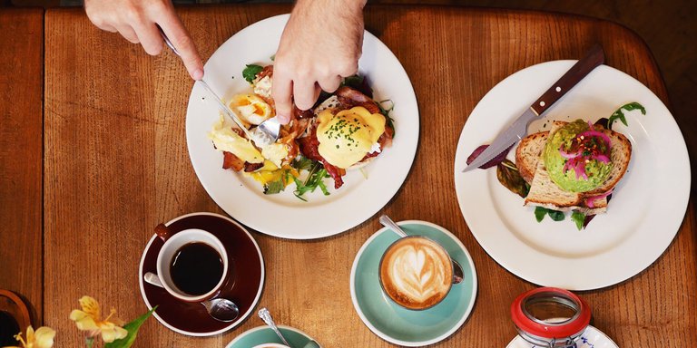 En översiktsbild på ett par personer som äter brunch tillsammans, en svart kaffe och en kaffe latte står på bordet tillsammans med en Eggs Benedict och en tallrik med Avokado-toast