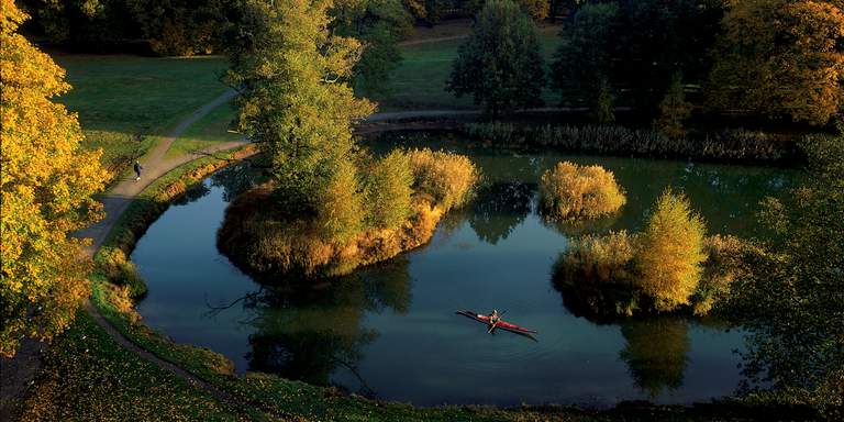 En fågelvy över Nationalstadsparken I Stockholm. En man promenerar med sin runt runt en damm, omgiven av träd med gulnande löv. Världens första nationalstadspark är en grön lunga som löper i en över tio kilometer lång båge kring och genom staden. Parklandskapet har kontakt med skogarna utanför vilket gör artrikedomen anmärkningsvärd. Här kan du möta rådjur och hare, rent av räv och älg, och sikta ovanliga fåglar, fjärilar och insekter.