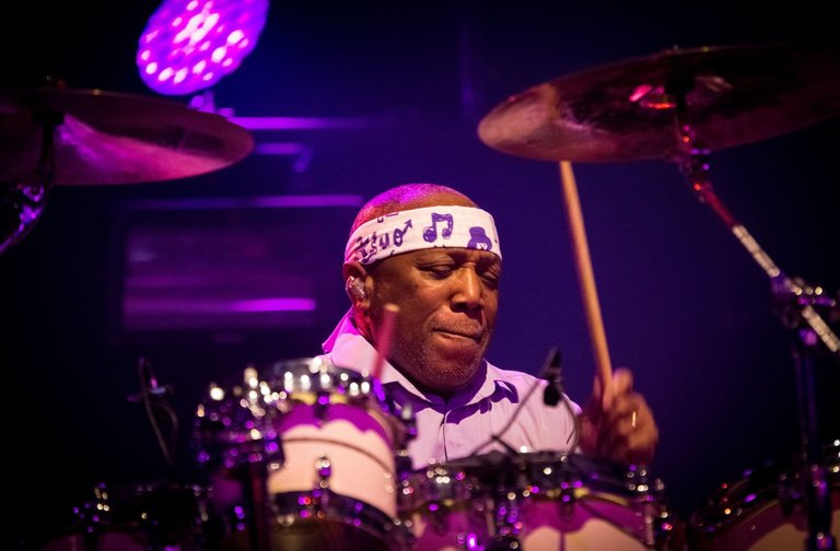 Trummisen Billy Cobham, spelar intensivt bakom ett trumset på en scen upplyst av lila och rosa scenljus. Han bär en vit pannband med noter och håller trumpinnar i rörelse. Bilden förmedlar energi, rytm och livekänsla.