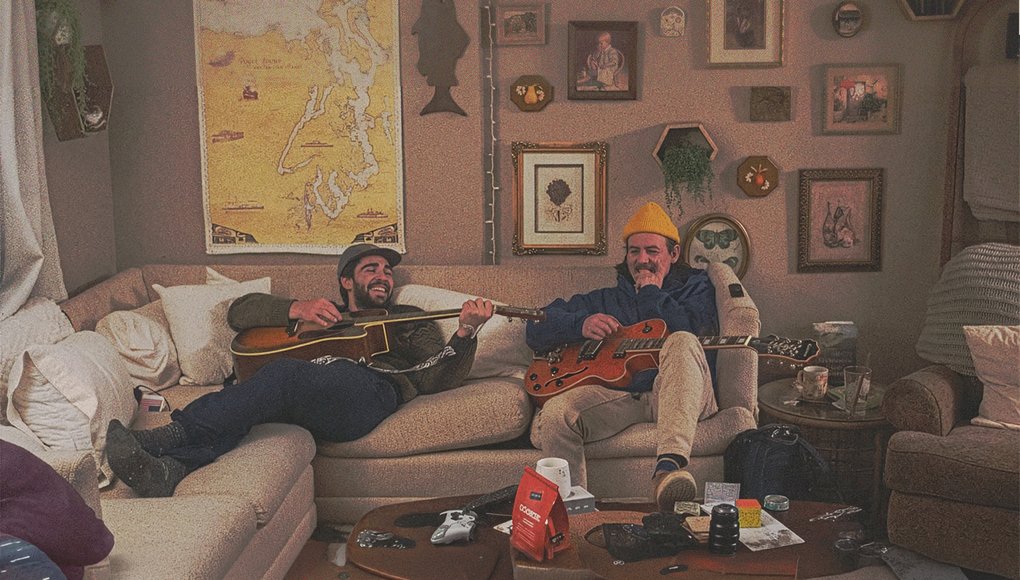 Två män sitter bekvämt på en soffa och spelar akustisk gitarr respektive elgitarr i ett rum med många prydnadsföremål och kartor på väggen.