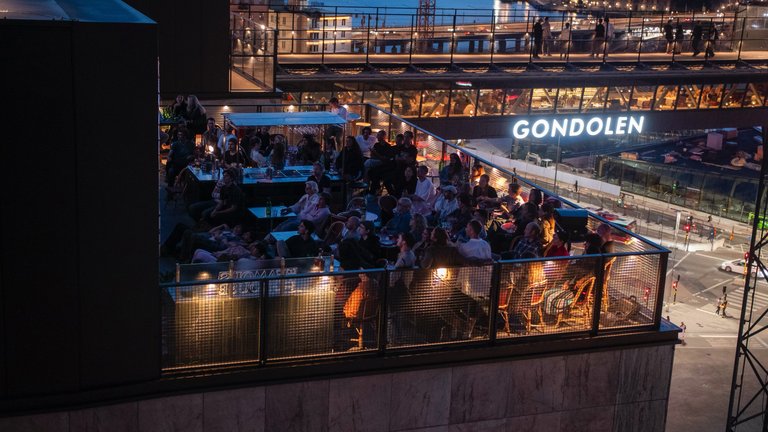 Kvällsscen på en takterrass i Slussen, Stockholm, där gäster sitter samlade med utsikt över stadsljusen och Gondolen i bakgrunden.