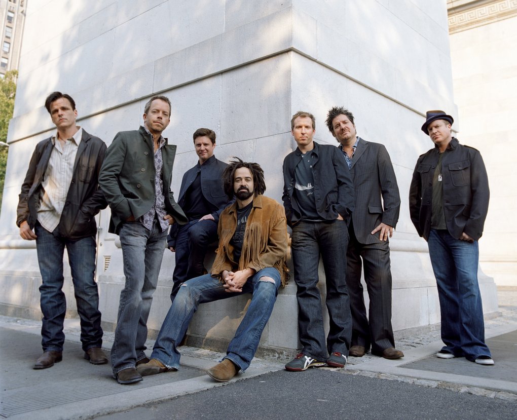 Counting Crows står och sitter lutade mot en stor vit stenbyggnad utomhus. De är klädda i vardagliga och halvt formella kläder Gruppen poserar avslappnat i rad.