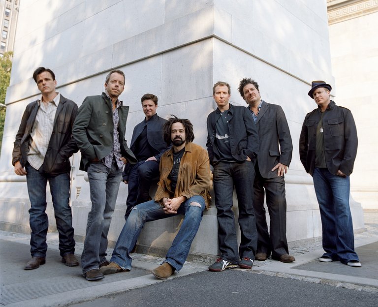 Counting Crows står och sitter lutade mot en stor vit stenbyggnad utomhus. De är klädda i vardagliga och halvt formella kläder Gruppen poserar avslappnat i rad.