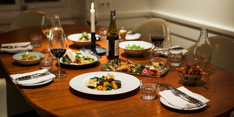  Ett ovalt bord star dukat, det ser nästan ut som man är hemma hos någon och en familj ska strax sätta sig ner och äta middag tillsammans. En flaska rött vin står på bordet och ett ljus är tänt. 