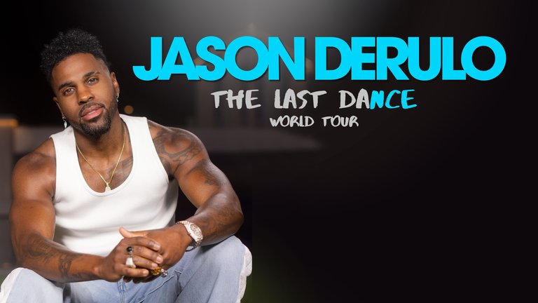 En tatuerad man med prydligt skägg, guldhalsband och vitt linne sitter för sig själv i ett mörkt rum.