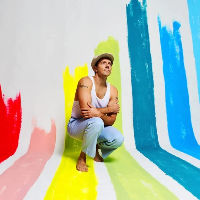 Jason Mraz i vitt linne och ljusblå byxor sitter på huk barfota, omgiven av breda färgstreck i rött, rosa, gult, grönt, turkos och blått som löper från vägg till golv. Han bär keps och ser tankfullt uppåt. Bakgrunden är vit med färgstarka kontraster.