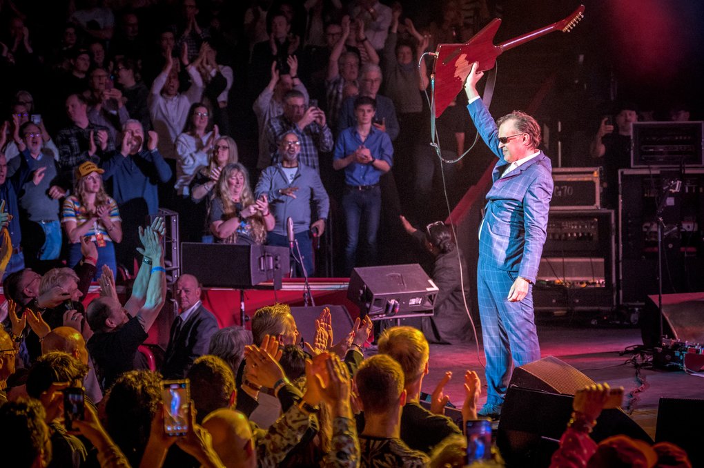 On a stage stands a man in a blue suit, holding up his electric guitar while surrounded by a crowd of adoring fans.