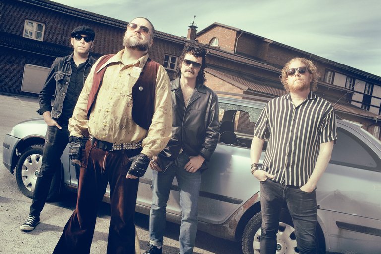 De fyra medlemmarna i Korslagda, iklädda solglasgögon står och poserar vid en silvrig kombi med rostfläckar.