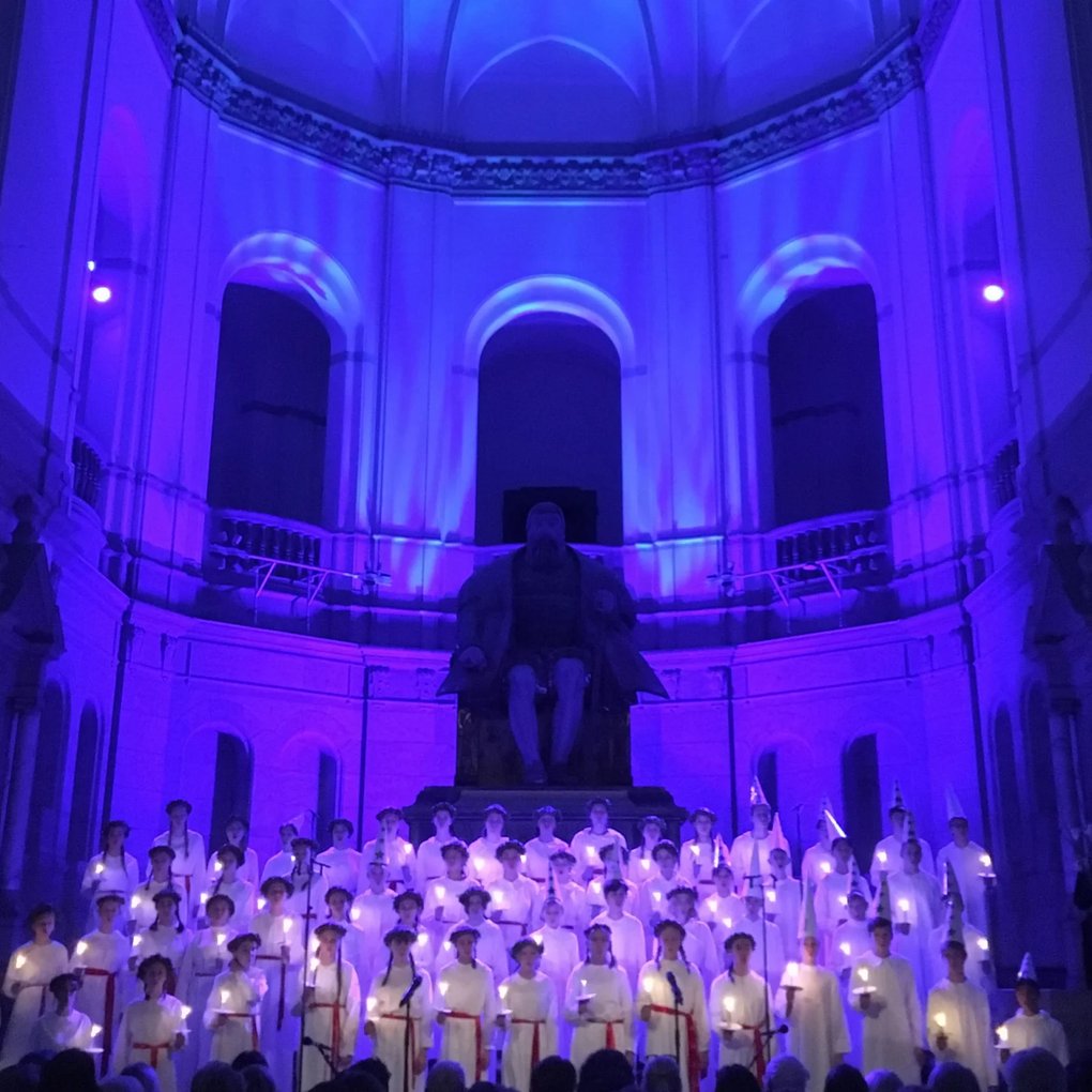 Traditionell Luciakör framför en stor staty av Gustav Vasa i en stor sal.