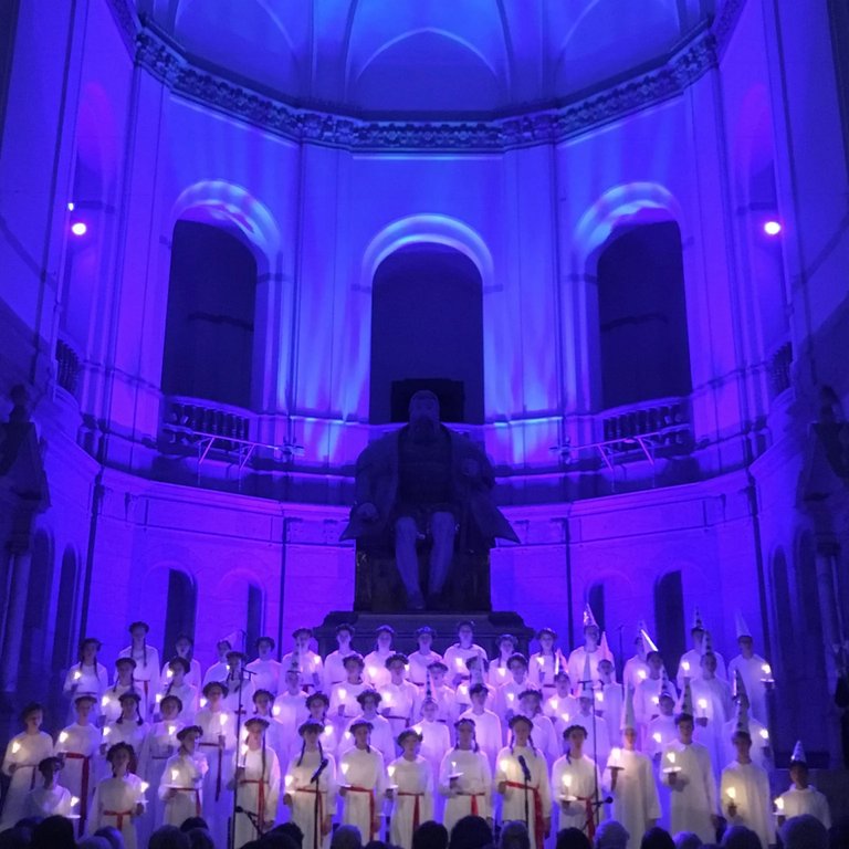 Traditionell Luciakör framför en stor staty av Gustav Vasa i en stor sal.