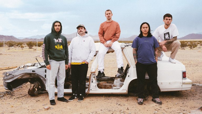 De fem medlemmarna i Militarie Gun poserar vid en urblåst bilkaross.