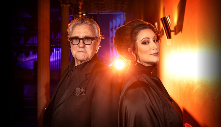 Tommy Körberg och Lisa Nilsson står rygg mot rygg i dramatisk belysning. Körberg bär svarta kläder och glasögon, medan Nilsson har elegant uppsatt hår och en svart dräkt. Ljuset bakom dem skapar en teatralisk och stämningsfull effekt.
