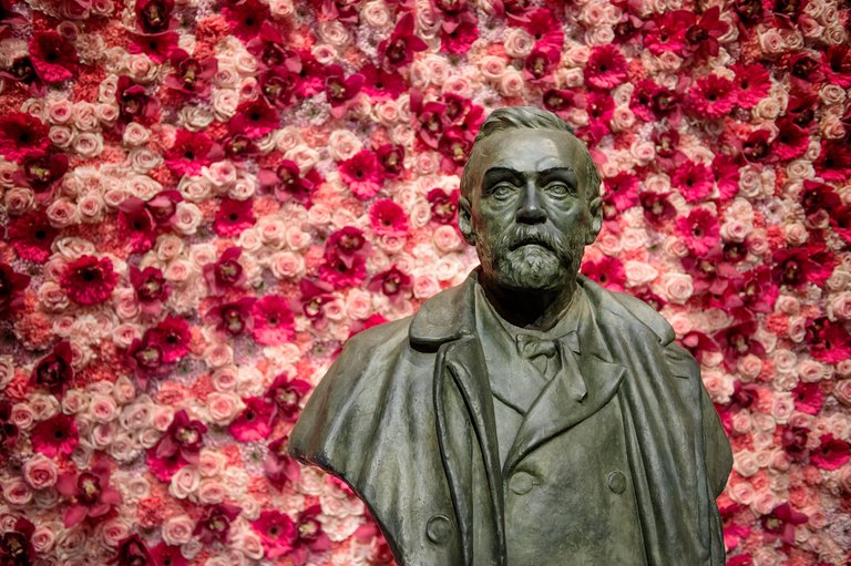 Bronze bust of a bearded Alfred Nobel in a coat against a wall of pink and red flowers.