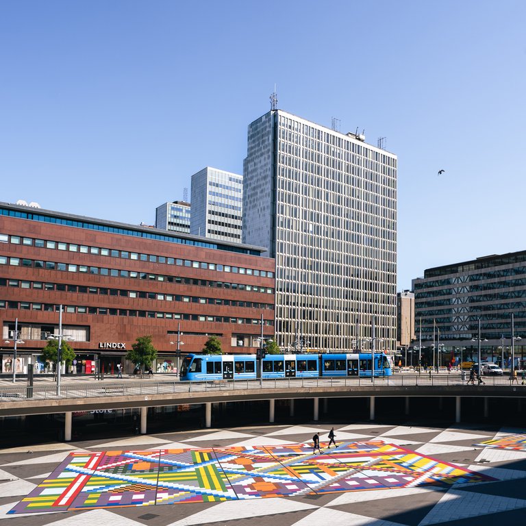 Arkitektur i Stockholm. Sergels torg i centrala Stockholm. På bilden syns skyskrapor, en spårvagn och torgets svartvita gatstenar. Gatstenarna har täckts med en färggrann matta.