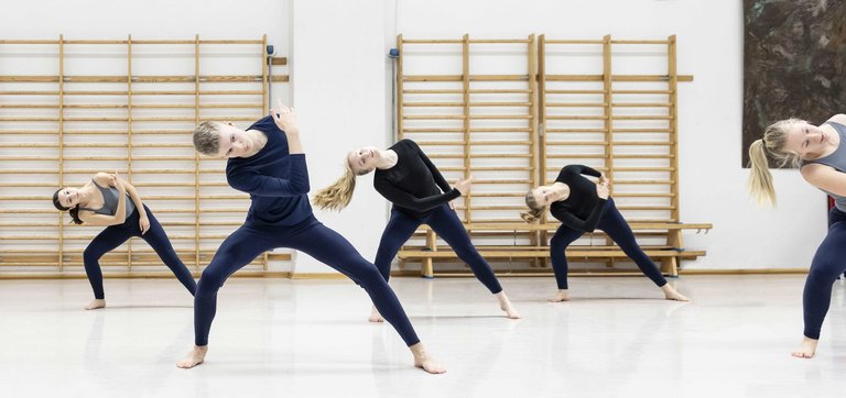 Grupp av samtida dansare i studio lutar sidledes i synk, barfota i mörkblå kläder framför ribbstolar.