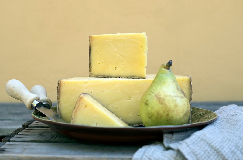 Tre bitar hårdost staplade på ett fat tillsammans med ett päron och en ostkniv på ett rustikt träbord.