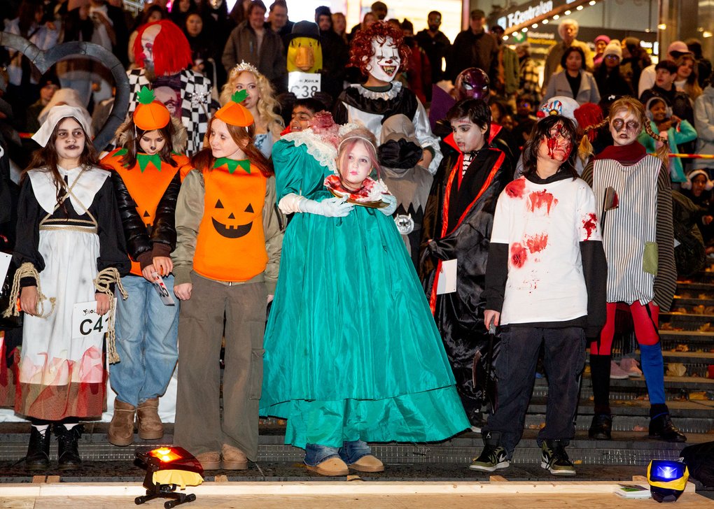 Gruppen poserar i olika Halloween-kostymer på en scen med publik i bakgrunden. Kostymerna inkluderar jack-o'-lantern-motiv, blodfläckar, teaterliknande kläder och skräckmasker.